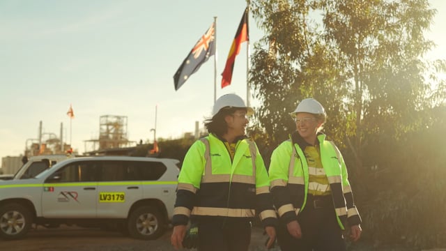 Two workers in high-visibility jackets and hard hats walking past a site vehicle, flags flying behind