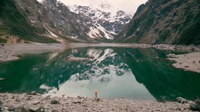 Lone figure on the stony shore of a still alpine lake reflecting snow-capped peaks