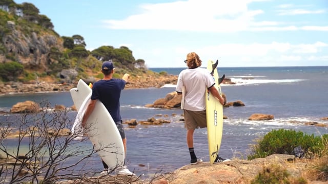 Two people carrying surfboards across a rocky headland above a bay