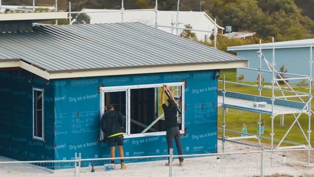 Two people fitting a window into a house wrapped in blue building wrap beside scaffolding