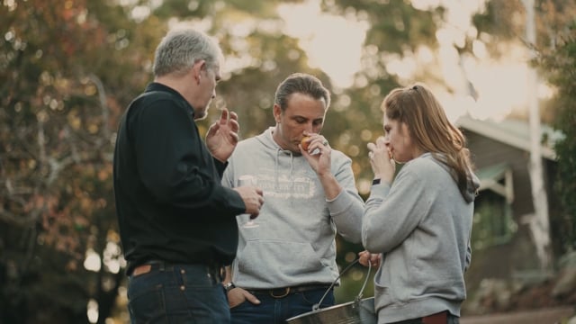 Three people tasting food and drink together outdoors under trees